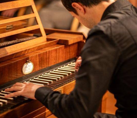 Ein Mann in einem schwarzen Hemd spielt ein altes Klavier mit einem Holzdeckel und Messingtasten vor einem sitzenden Publikum.