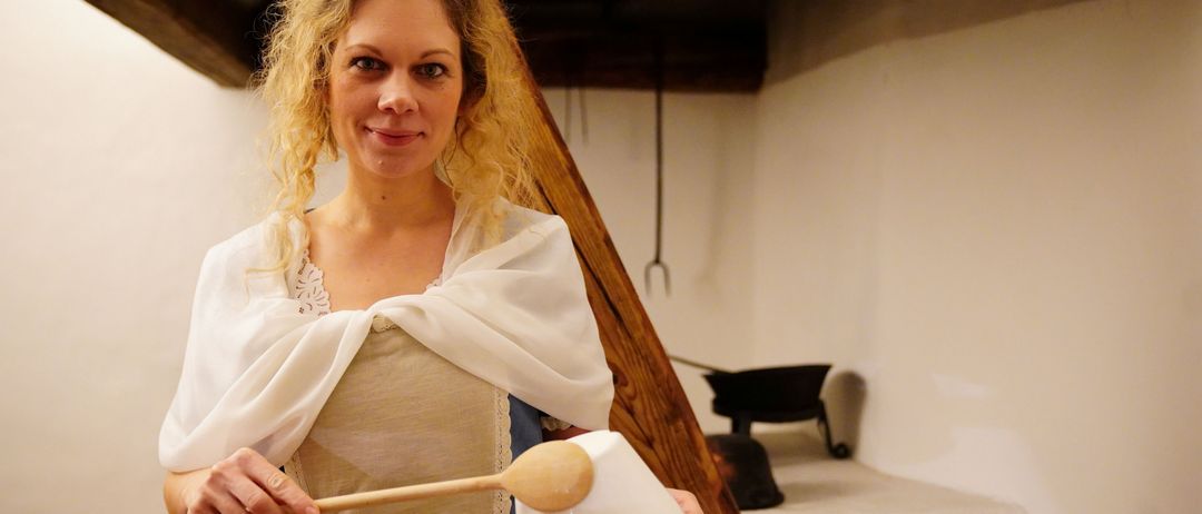 Eine Frau mit einem weißen Umhang und einem blauen Kleid lächelt und hält einen Holzlöffel und eine weiße Schüssel. Sie scheint sich in einem Raum mit Holzbalken und weißen Wänden zu befinden.