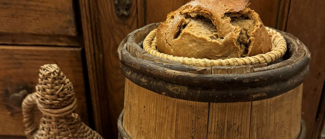 Ein rustikaler Holzfass mit einem geflochtenen Korbdeckel steht auf einem gefliesten Boden und enthält ein Brot. Daneben steht eine geflochtene Flasche.