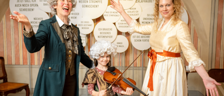Drei Personen in historischen Kostümen stehen vor einer Wand mit deutschem Text. Die Frau in der Mitte hält eine Geige, und die beiden anderen gestikulieren lebhaft.