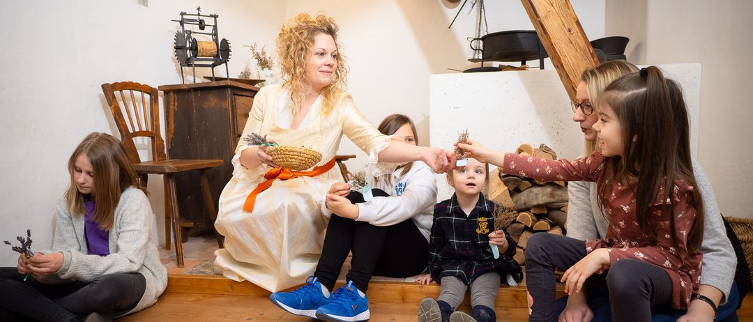 Eine Frau in einem Kleid hält einen Korb, während Kinder um sie herum sitzen, einer hält ein kleines Objekt. Der Raum ist rustikal mit Holzbalken und Möbeln.