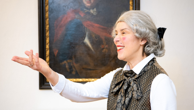 Eine ältere Frau, in einem Kostüm aus vergangenen Zeiten gekleidet, steht vor einem Porträtgemälde und gestikuliert lächelnd mit den Händen.
