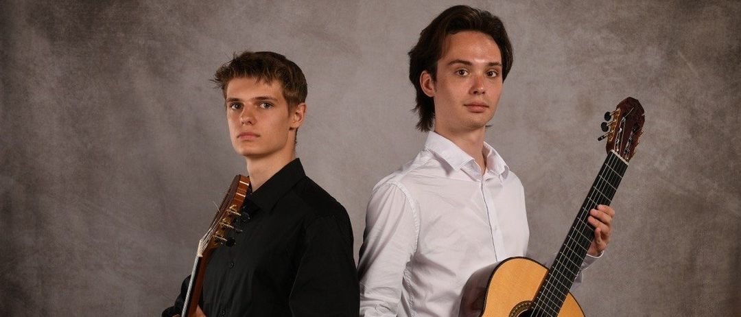 Zwei junge Männer stehen nebeneinander und halten jeweils eine klassische Gitarre. Der Mann auf der linken Seite trägt ein schwarzes Shirt, der Mann auf der rechten Seite ein weißes. Sie schauen beide nach vorne, möglicherweise für ein Foto posierend.