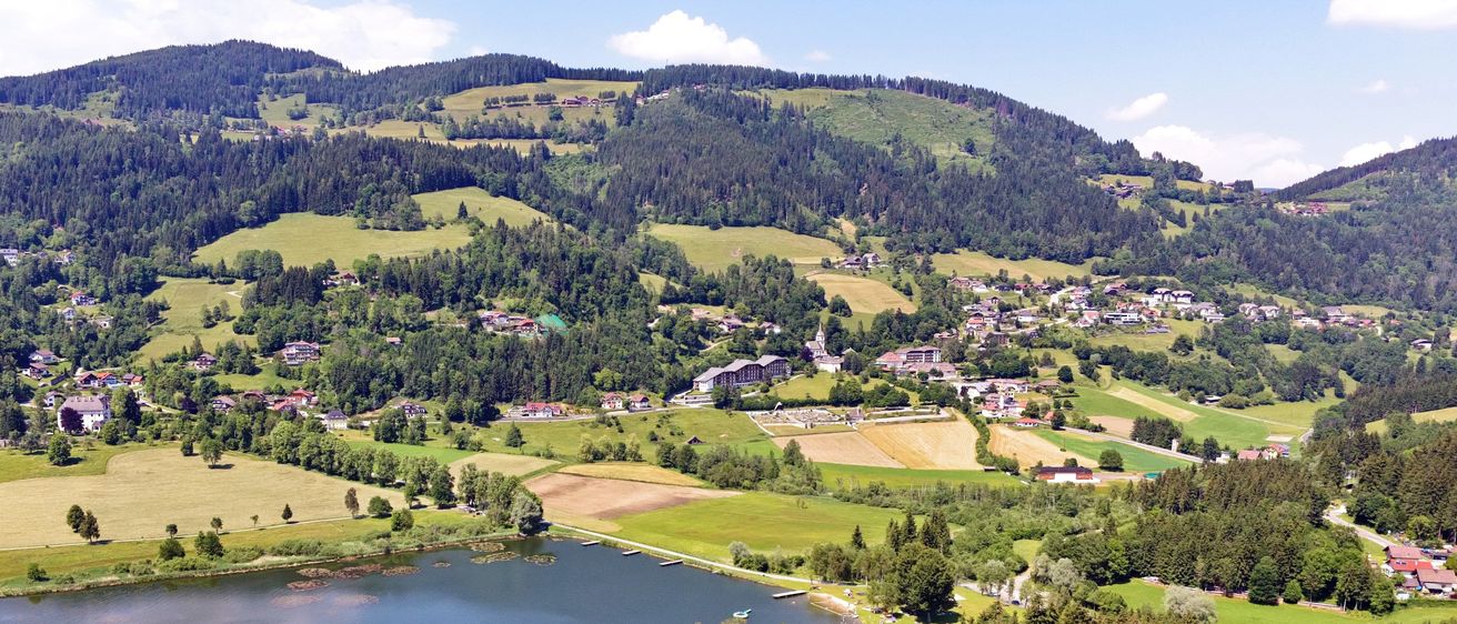 Ein Luftbild einer ländlichen Landschaft mit einem See, umgeben von grünen Feldern und Wäldern. Der Himmel ist blau mit einigen Wolken.