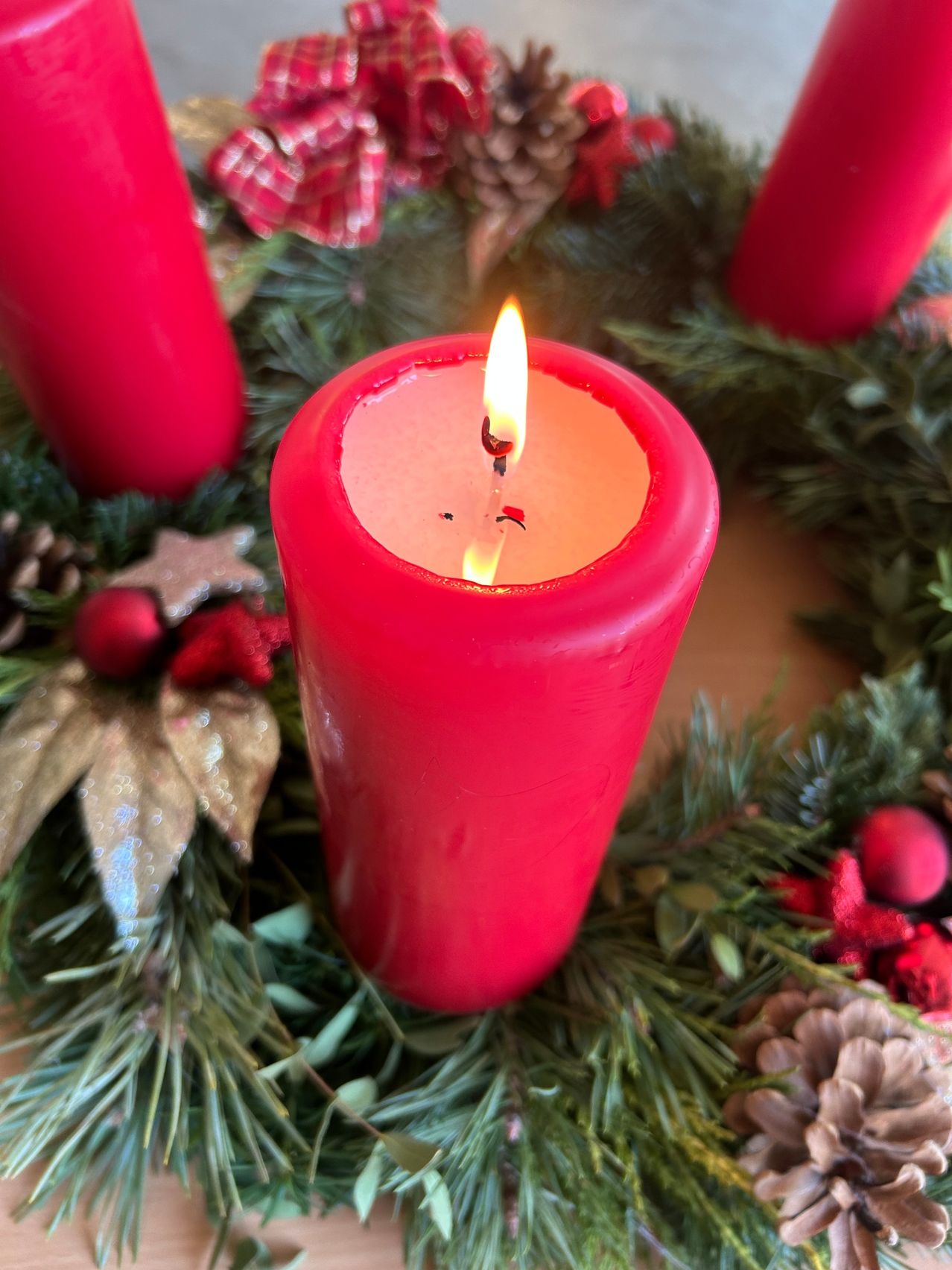 Eine rote Kerze ist in der Mitte eines Kranzes angezündet, umgeben von Tannenzapfen und Ornamenten. Die Kerze ist teilweise geschmolzen, mit einer sanft leuchtenden Flamme.