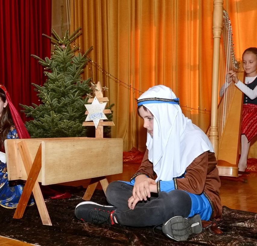 Ein junger Junge, der als biblische Figur gekleidet ist, sitzt neben einer hölzernen Krippe mit einem Weihnachtsbaum und einem Stern darauf. Hinter ihm spielt ein junges Mädchen Harfe.