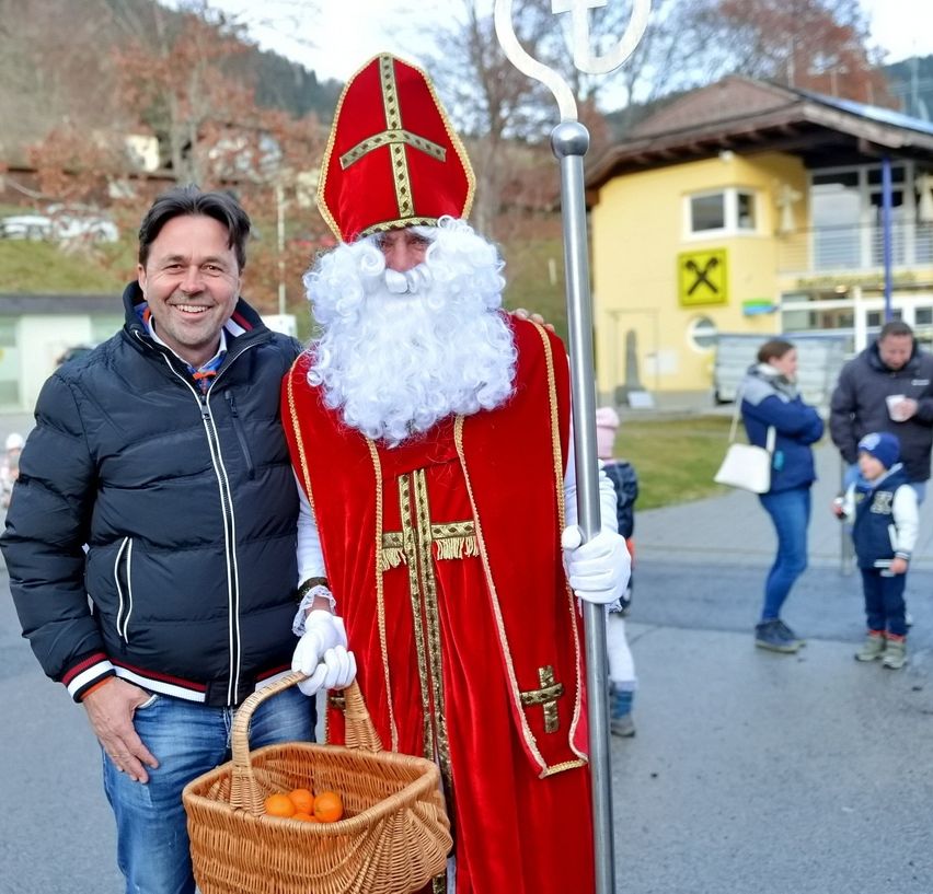 Ein Mann, der als Weihnachtsmann verkleidet ist, hält einen Korb mit Orangen in der Hand und steht neben einem Mann in einer schwarzen Jacke. Sie sind draußen in der Nähe eines Gebäudes mit gelber Fassade.