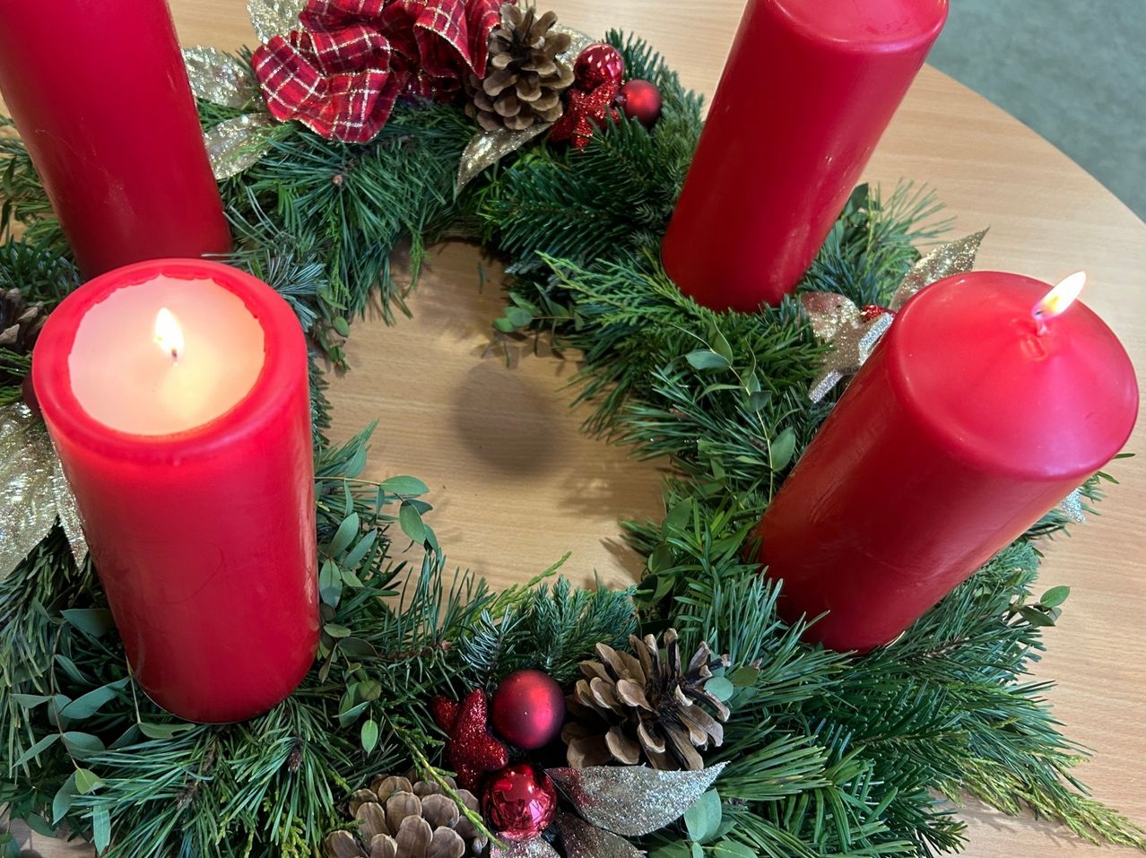 Ein Kranz mit roten Kerzen auf einem Holztisch. Grünes Laub, silberne Bänder und Tannenzapfen sind ebenfalls vorhanden. Eine Kerze ist angezündet.