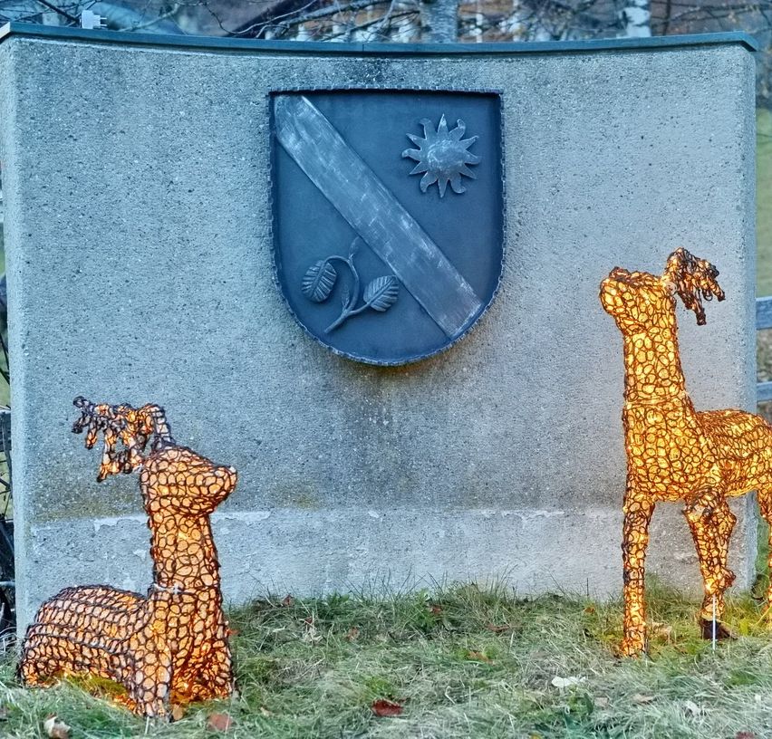 Eine Betonwand mit einem angebrachten Schild, das eine Sonne und Blätter zeigt. Zwei beleuchtete Rentierstatuen stehen vor der Wand auf dem Rasen.
