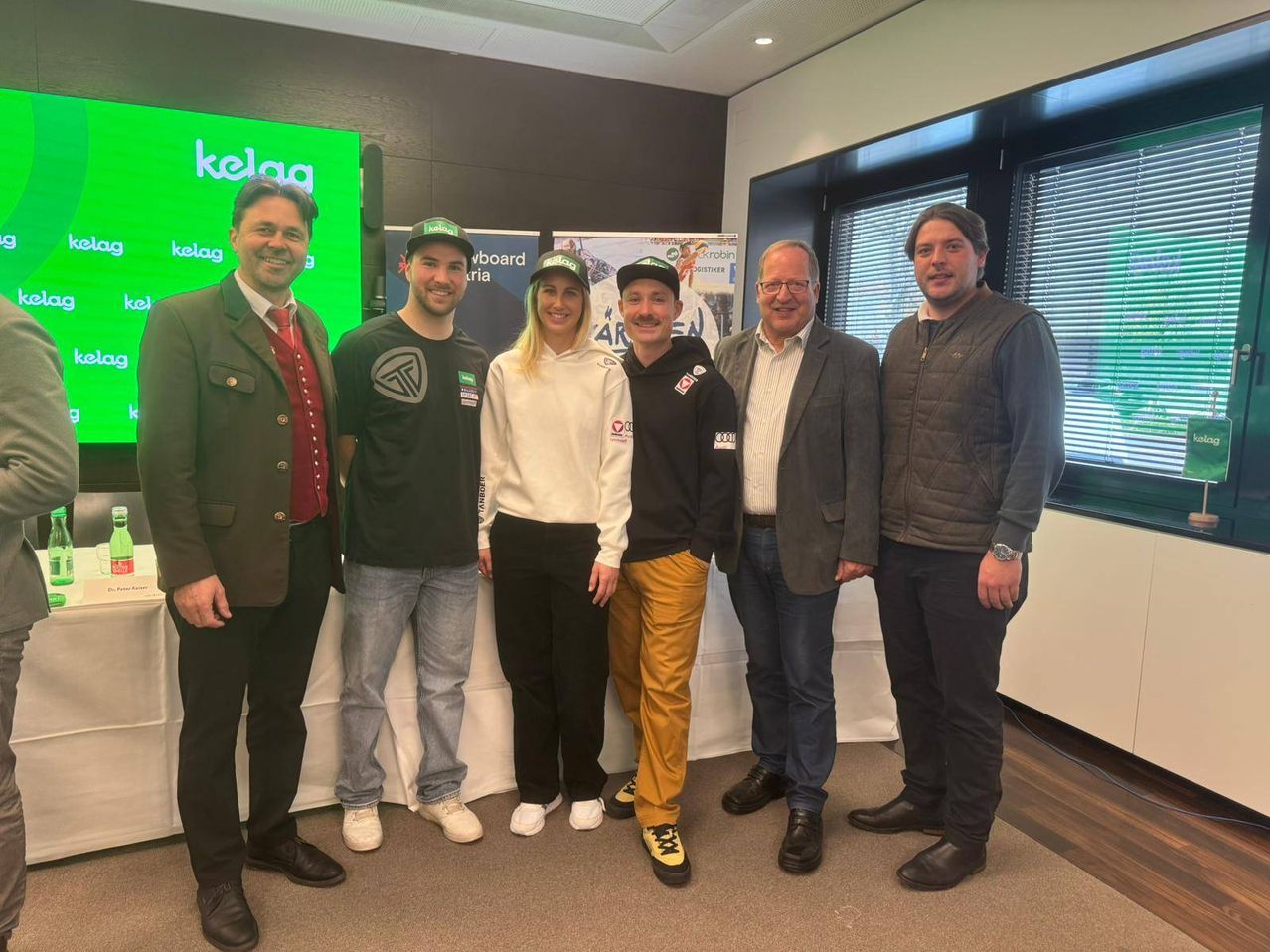 Fünf Personen stehen eng zusammen vor einer grünen Wand mit einem KELA-Logo. Die Gruppe besteht aus einem Mann mit Hut, einer Frau in einer weißen Hoodie und einem Mann in einem schwarzen Jackett. Sie lächeln und scheinen für ein Foto zu posieren.