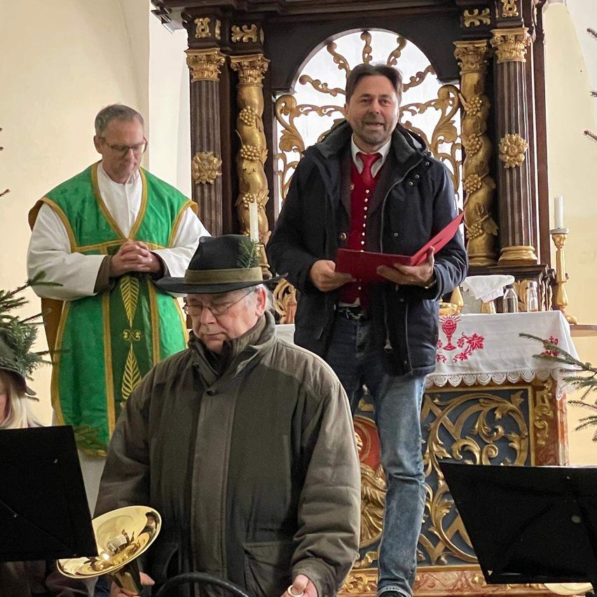 Ein Mann mit einer roten Mappe steht vor einem Altar, hinter ihm ein Priester in grünen Gewändern. Ein weiterer Mann in einem Hut spielt eine goldene Trompete.