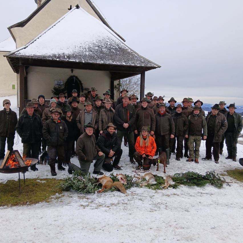 Eine Gruppe von Menschen, überwiegend Männer, stehen und knien in einem verschneiten Bereich vor einer Kapelle. Sie tragen alle Winterkleidung und Hüte. Einige von ihnen halten Hunde. Es gibt Tannenzweige auf dem Boden und ein Lagerfeuer in der linken Ecke.