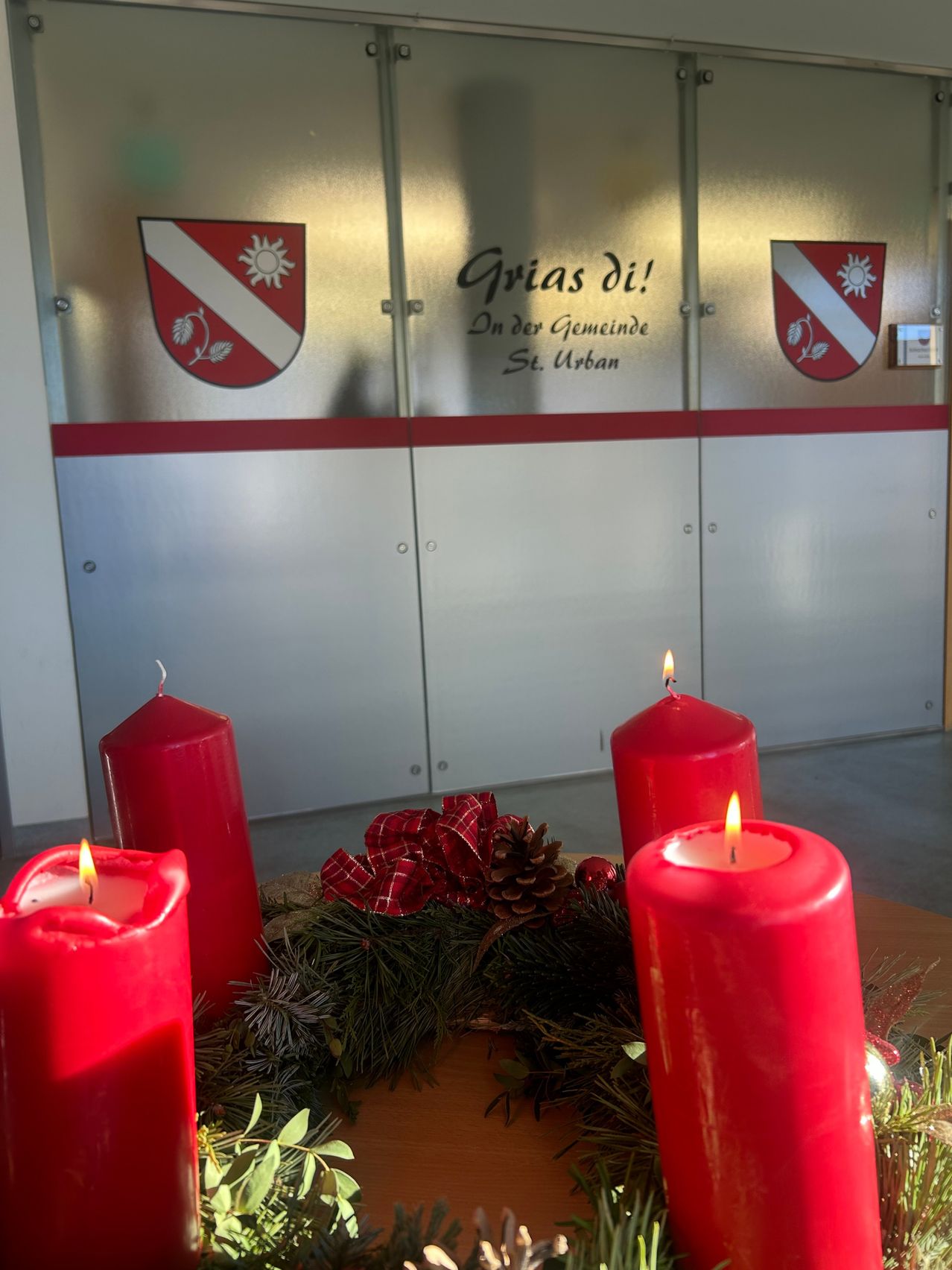 Ein Raum mit festlicher Dekoration, bestehend aus vier roten Kerzen und einem Kranz mit Tannenzapfen und Schleifen. Hinter den Kerzen befindet sich eine metallische Wand mit rotem Rand und Logos.