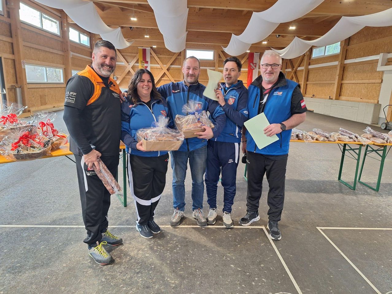 Fünf Personen in blauen Jacken und Turnschuhen stehen in einem Saal und halten eingewickelte Geschenke. Ein Mann hält ein Stück Papier und ein Stück Essen. Ein Tisch mit Essen steht hinter ihnen.