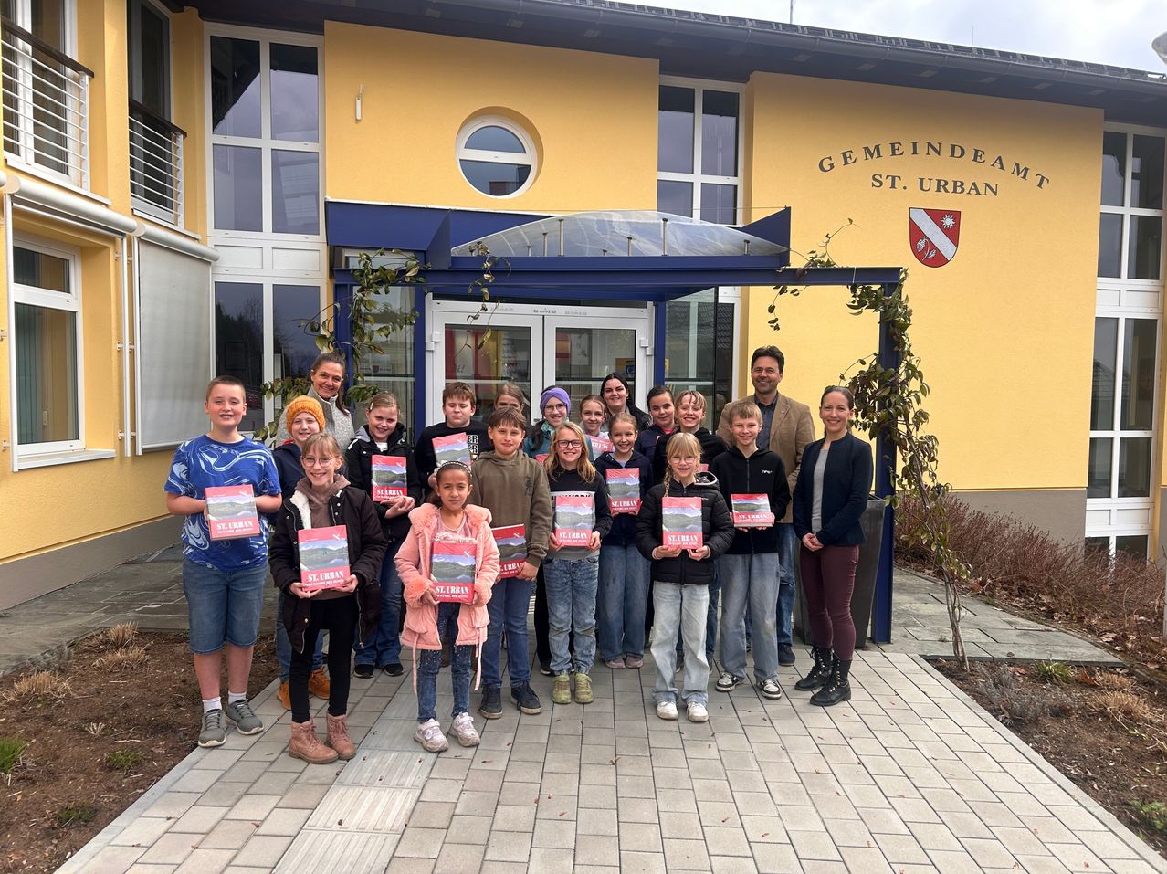 Eine Gruppe von Kindern und Erwachsenen posiert für ein Foto vor einem Gebäude mit der Aufschrift 'Gemeindeaam St. Urban'. Sie halten Bücher in den Händen.