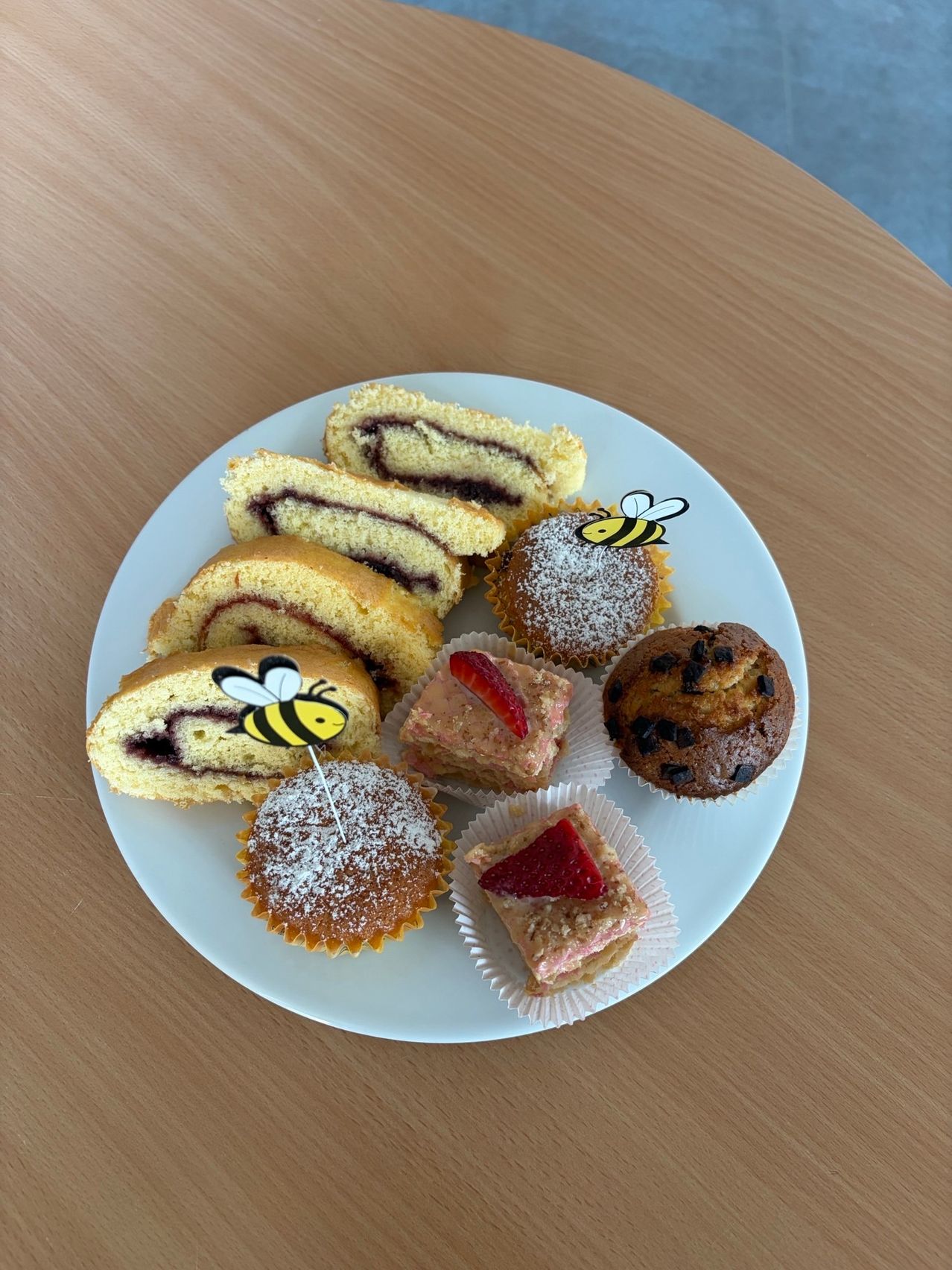 Ein weißer Teller auf einem Holztisch zeigt eine Auswahl an Desserts. Es gibt Erdbeertarte, Cupcakes und einen gerollten Kuchen mit Himbeerfüllung, alle mit Bienenaufklebern verziert.