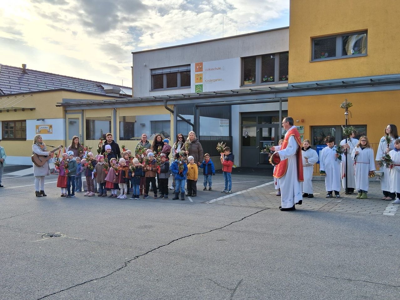 Eine Gruppe von Kindern mit Blumen steht vor einem Priester, mit einem Gebäude im Hintergrund.