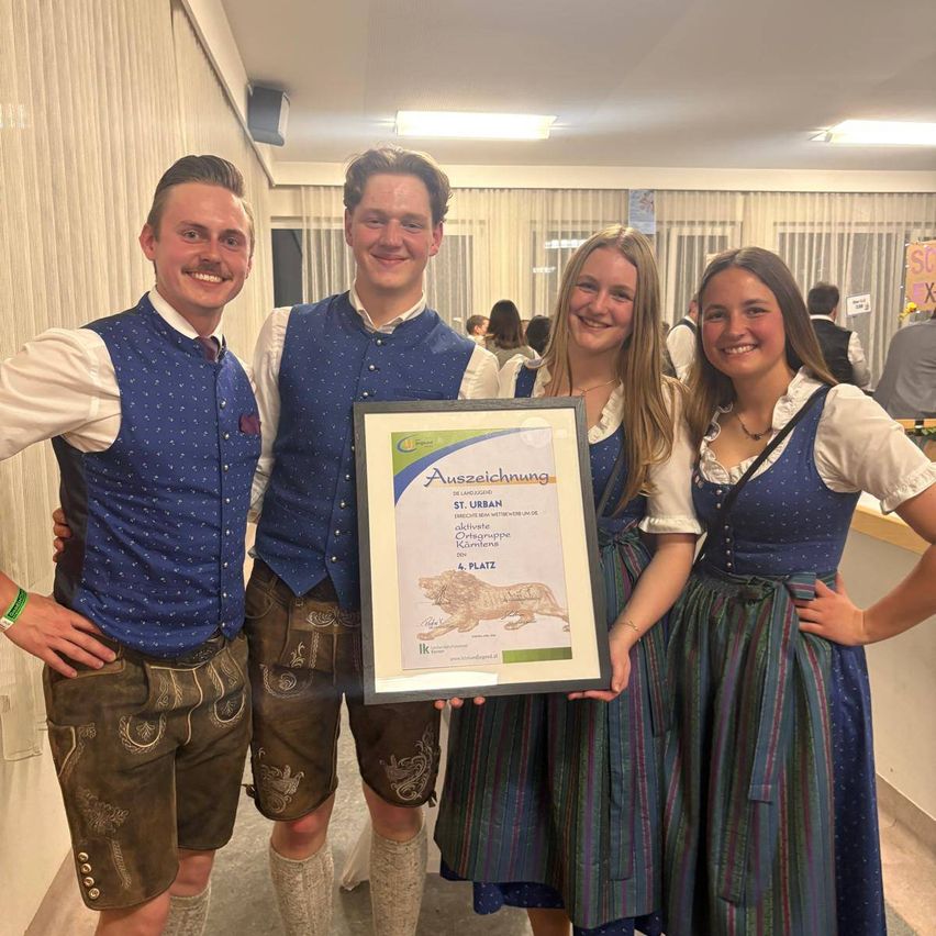 Vier Menschen lächeln und posieren für ein Foto. Zwei Männer und zwei Frauen tragen traditionelle deutsche Kleidung. Sie halten ein gerahmtes Zertifikat.