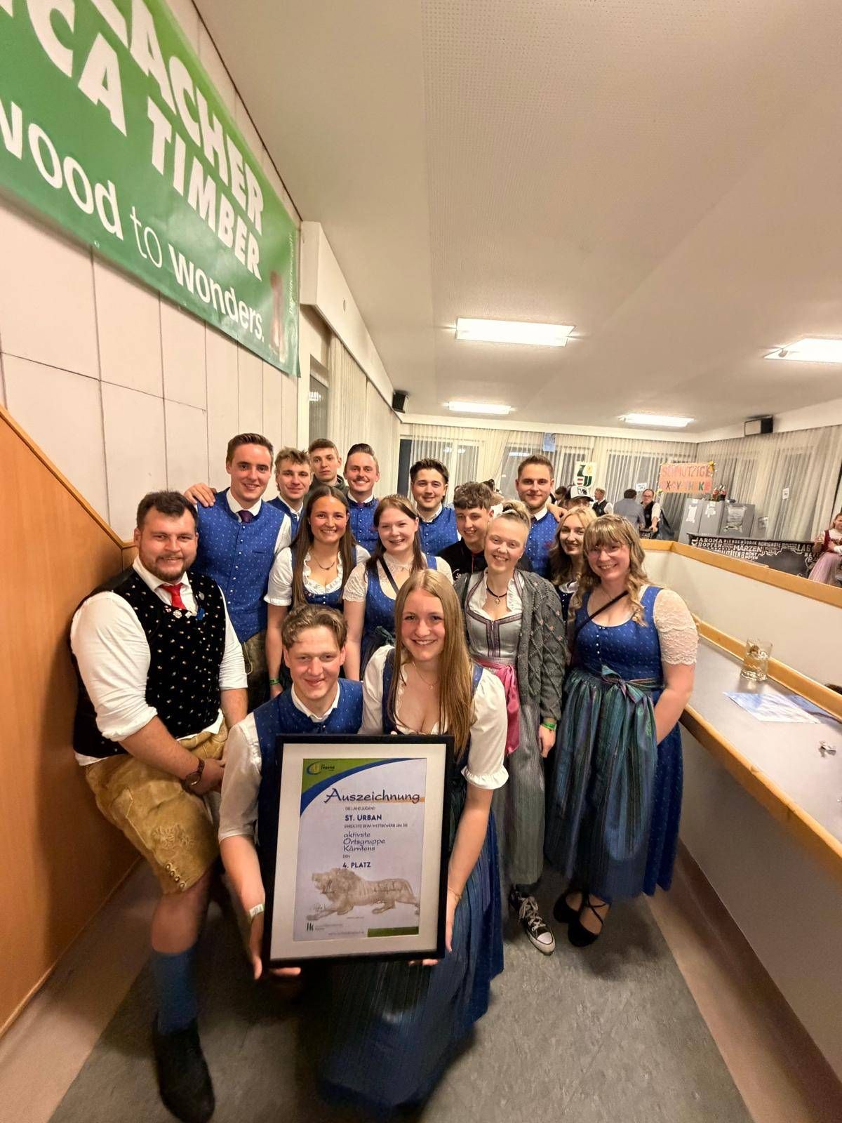 Eine Gruppe von Menschen in traditioneller Kleidung steht in einem Korridor und hält einen in Gold gerahmten Preis. Eine Frau in der Mitte lächelt, während sie den Rahmen hält, mit anderen lächelnden und posierenden Personen hinter ihr. Ein Banner mit Text und Logo hängt an der Wand.