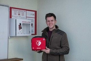 Ein Mann hält einen roten Philips-Defibrillator in einem Raum mit einer weißen Wand und einem roten Schild. Er trägt einen braunen Mantel und steht neben einer weißen Mikrowelle.