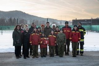 Eine Gruppe von Menschen in Winterkleidung steht in einer Reihe auf einem schneebedeckten Außengelände, mit Bergen und einem Gebäude im Hintergrund.