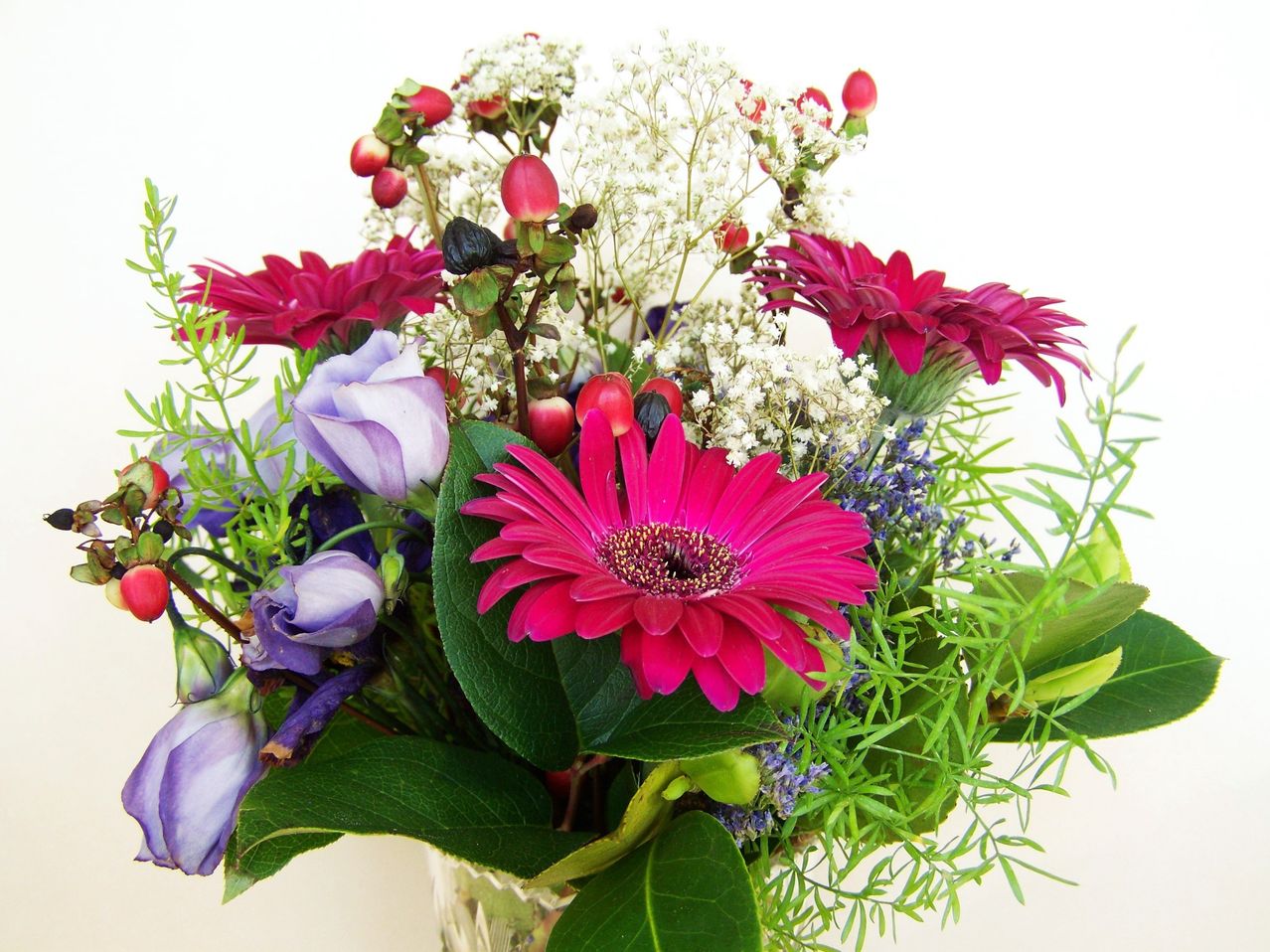 Eine farbenfrohe Blumenarrangement mit pinken Gerbera-Daisies, lila Rosen und verschiedenen Grünpflanzen. Vor einem weißen Hintergrund präsentieren die Blumen eine Mischung aus Farben und Texturen.