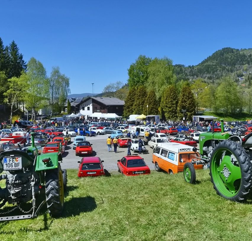 Ein großes Autotreffen unter einem klaren blauen Himmel, umgeben von Bäumen und Bergen, mit Traktoren, Zelten und einem Gebäude in der Ferne.