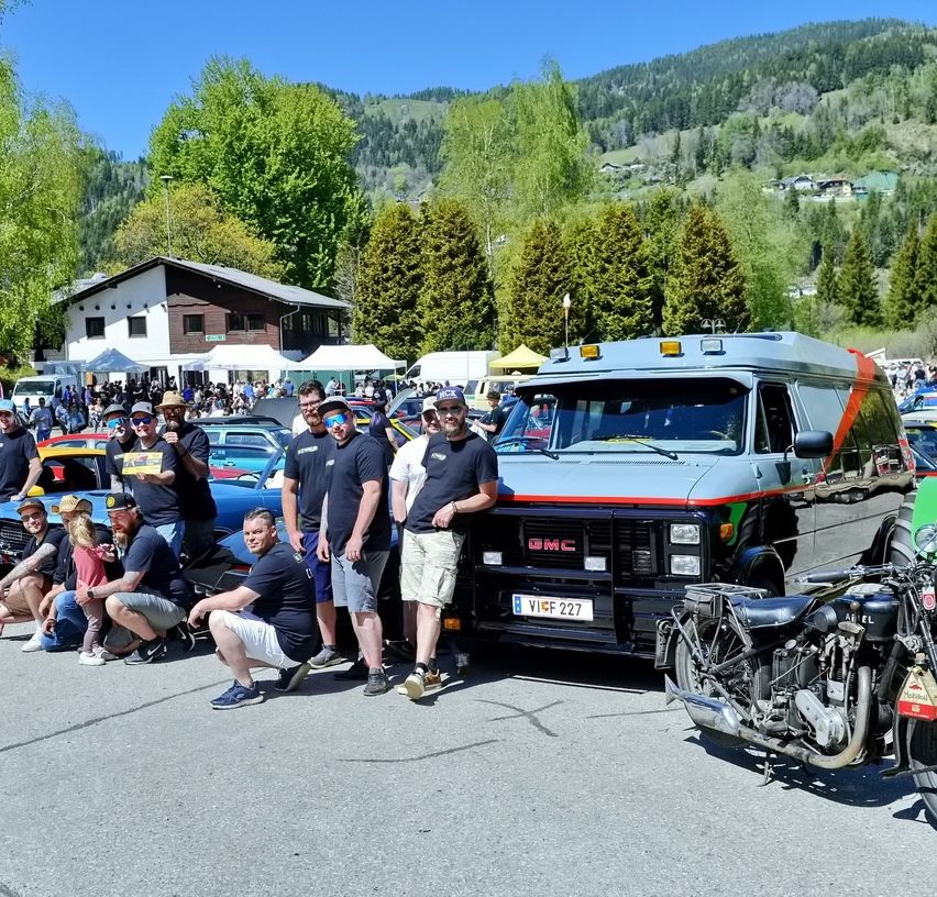 Eine Gruppe von Menschen steht vor einem GMC-Van und einem Motorrad auf einem Parkplatz. Der Van und das Motorrad sind in der Nähe eines Berges geparkt.