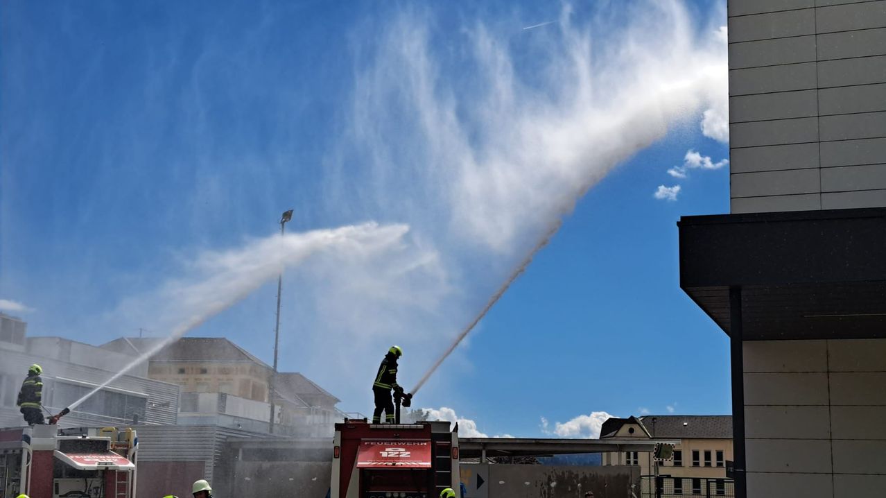 Ein Feuerwehrmann steht auf der Spitze eines Feuerwehrwagens und sprüht Wasser aus einer Düse. Das Wasser spritzt nach oben in den Himmel. Im Hintergrund sind Gebäude zu sehen.
