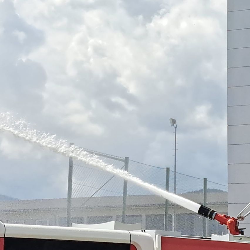 Ein Feuerwehrwagen sprüht Wasser durch einen Strahl, der auf einen Zaun gerichtet ist, unter einem bewölkten Himmel.