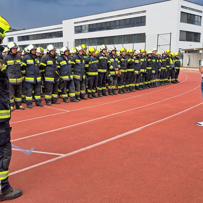 Eine Reihe von Feuerwehrleuten steht auf einer Laufbahn, alle tragen gelbe und schwarze Uniformen, Helme und Stiefel. Sie befinden sich wahrscheinlich in einer Trainingseinheit. Ein weißes Gebäude steht hinter ihnen.