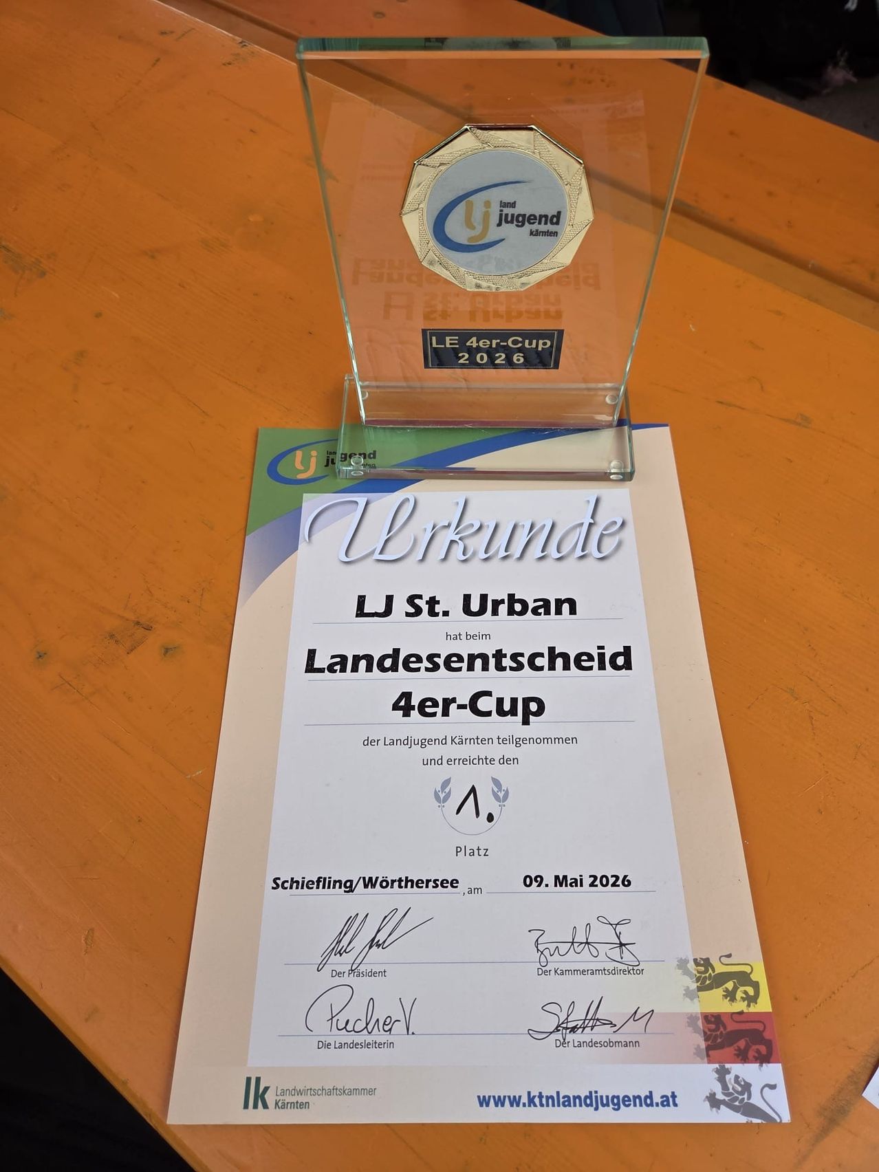 Ein durchsichtiger Display-Schrank hält eine kreisförmige Auszeichnung, beschriftet mit 'LE 4er-Cup 2026', auf einer orangefarbenen Oberfläche. Darunter steht ein Zertifikat mit 'Urkunde LJ St. Urban Landesentscheid 4er-Cup'. Es ist vom Präsidenten und dem Kammerpräsidenten unterzeichnet.