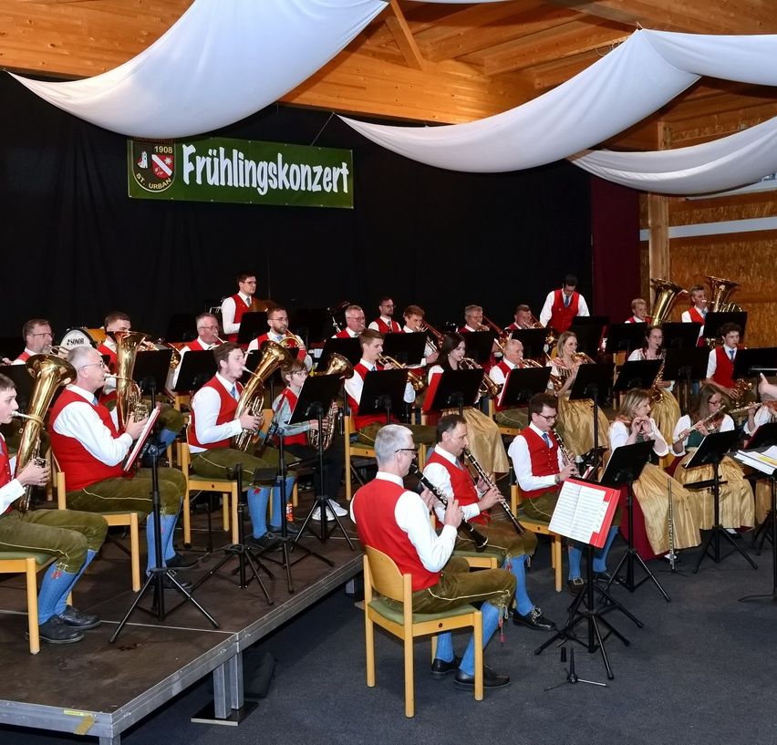 Ein Orchester spielt auf der Bühne mit einem Banner darüber, das Frühlingskonzert sagt. Sie spielen verschiedene Instrumente, darunter Trompeten und Posaunen.