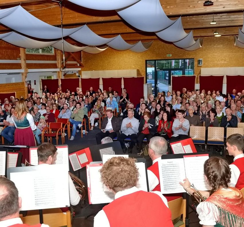 Ein großes Publikum versammelt sich in einem Saal, klatscht und beobachtet ein Blasorchester in roten Westen. Die Musiker stehen auf der Bühne mit Notenständern vor ihnen. Der Saal hat Holzbalken und rote Vorhänge.