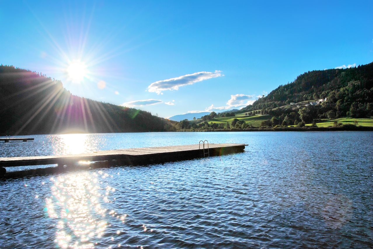 Ein ruhiger See mit einem hölzernen Steg, der sich ins Wasser erstreckt, umgeben von üppigem Grün und Bergen unter einem klaren blauen Himmel.