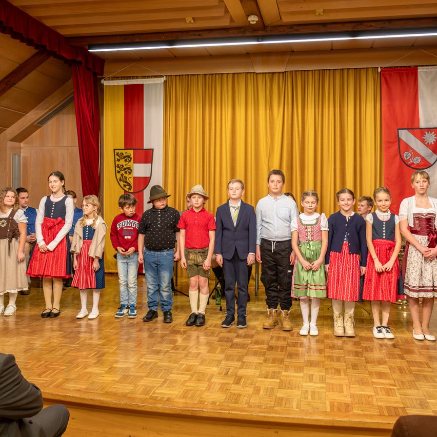 Eine Gruppe von Kindern und einigen Erwachsenen steht auf der Bühne in traditioneller Kleidung. Hinter ihnen befinden sich gelbe Vorhänge und Fahnen.