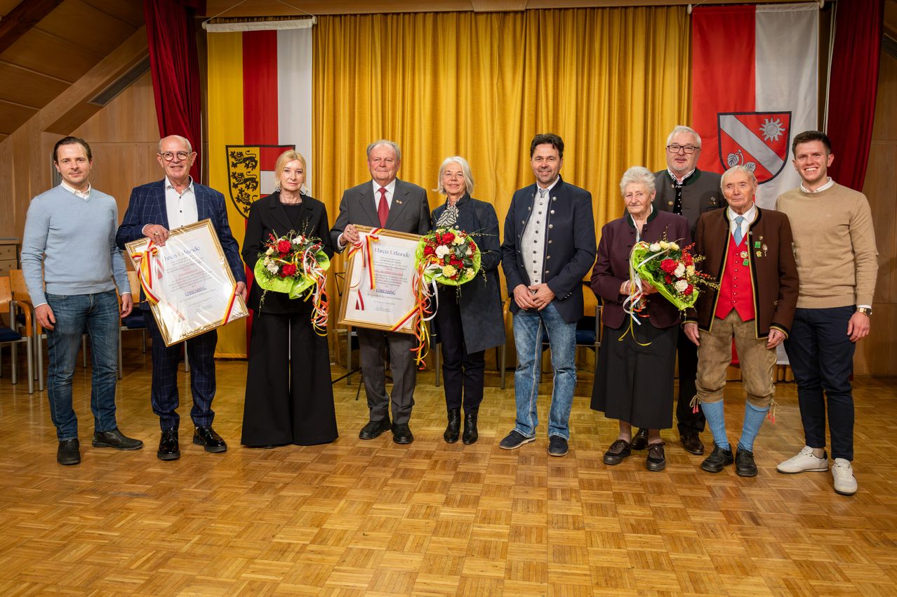 Eine Gruppe von Menschen steht auf einem Holzboden und hält Blumen und Urkunden. Sie scheinen eine Preisverleihung zu feiern.