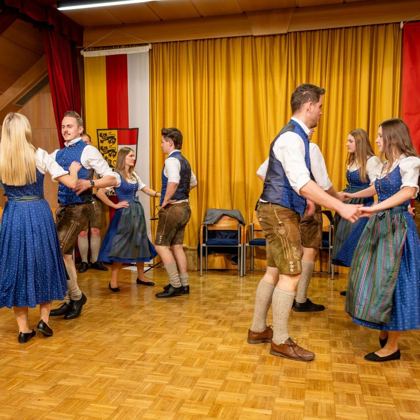 Eine Gruppe von Menschen in traditioneller deutscher Kleidung tanzt zusammen auf einem Holzboden. Hinter ihnen befinden sich gelbe Vorhänge und eine Flagge.