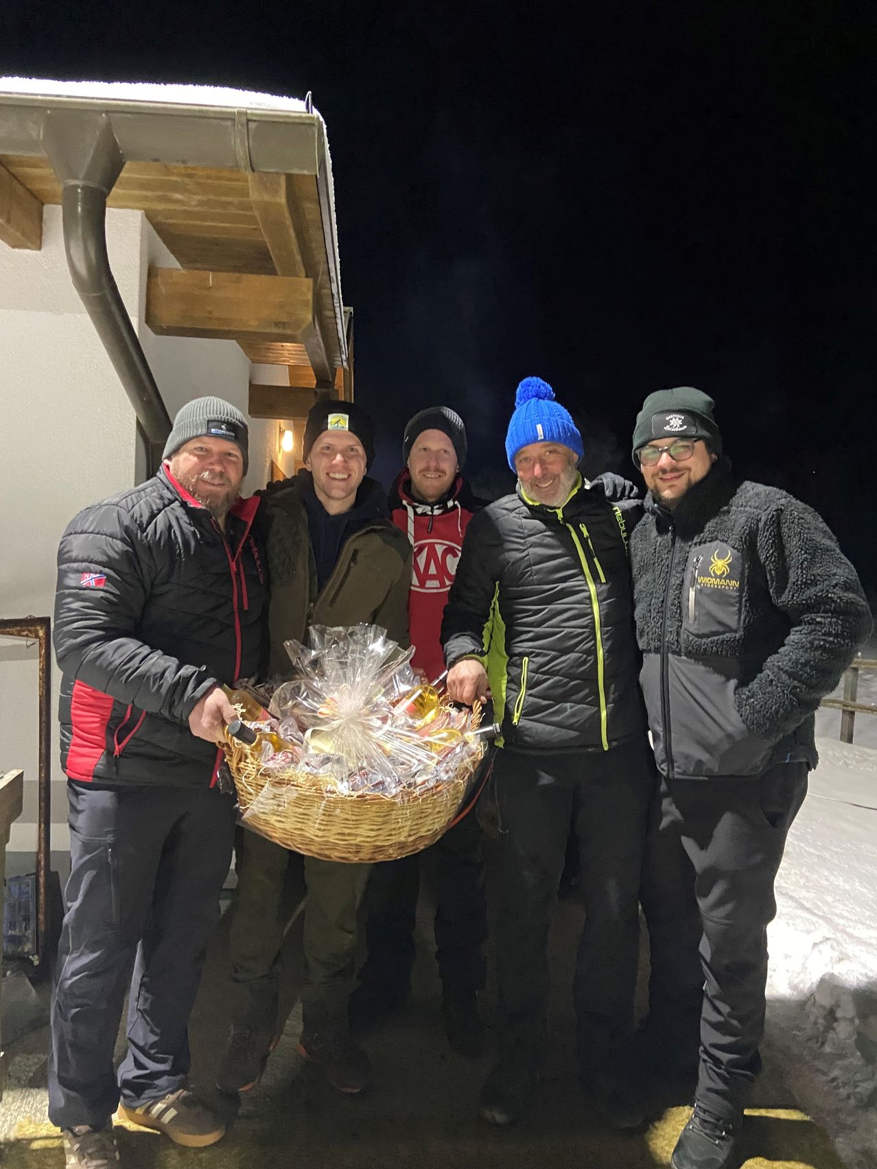 Fünf Männer stehen nachts im Freien und lächeln. Einer hält einen Korb voller verpackter Gegenstände. Sie tragen Winterjacken und Mützen. Hinter ihnen steht eine Holzstruktur.