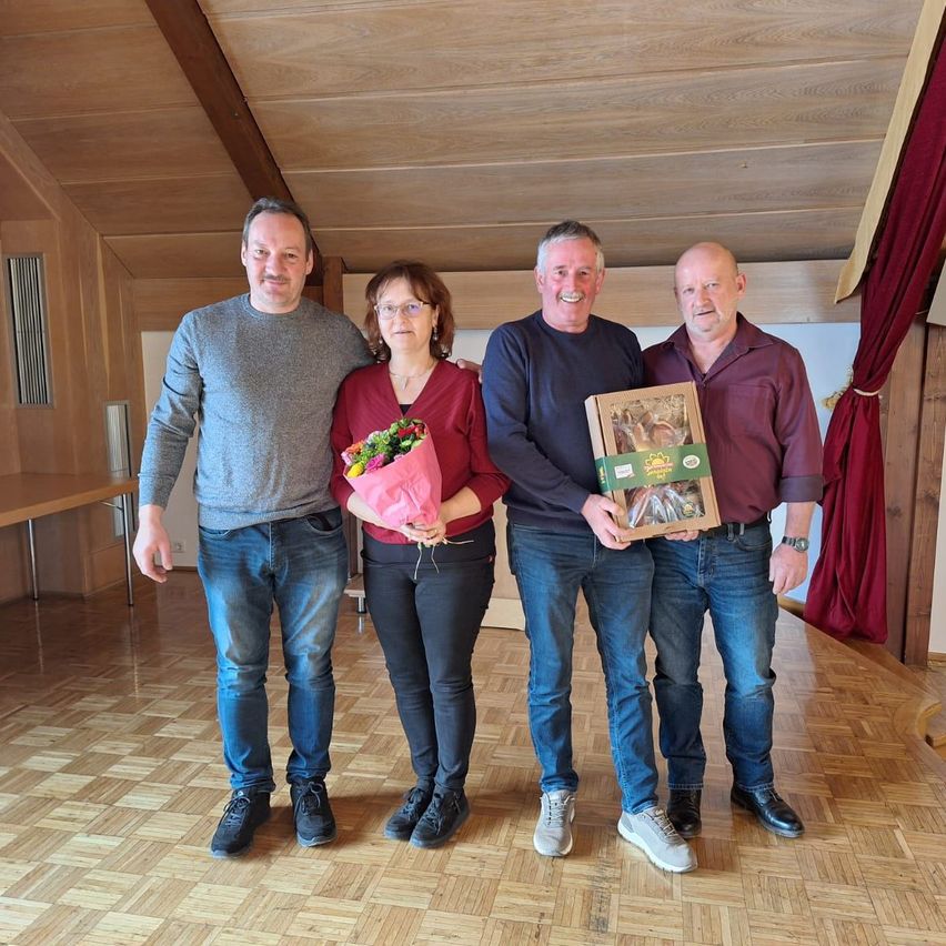 Vier Personen stehen lächelnd in einem Raum mit Holzböden. Ein Mann auf der linken Seite hält Blumen, ein anderer eine Box, und eine Frau steht zwischen ihnen.