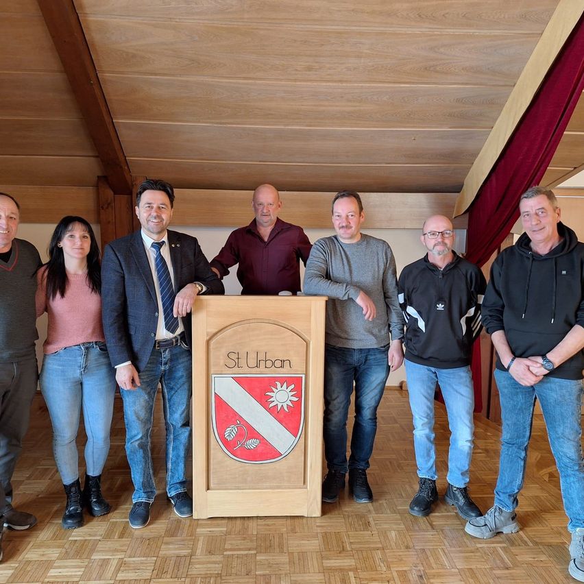 Sieben Personen stehen in einem Raum. Drei Männer stehen hinter einem Podium mit einem Logo und der Aufschrift 'St Urban'. Die Gruppe scheint für ein Foto zu posieren.