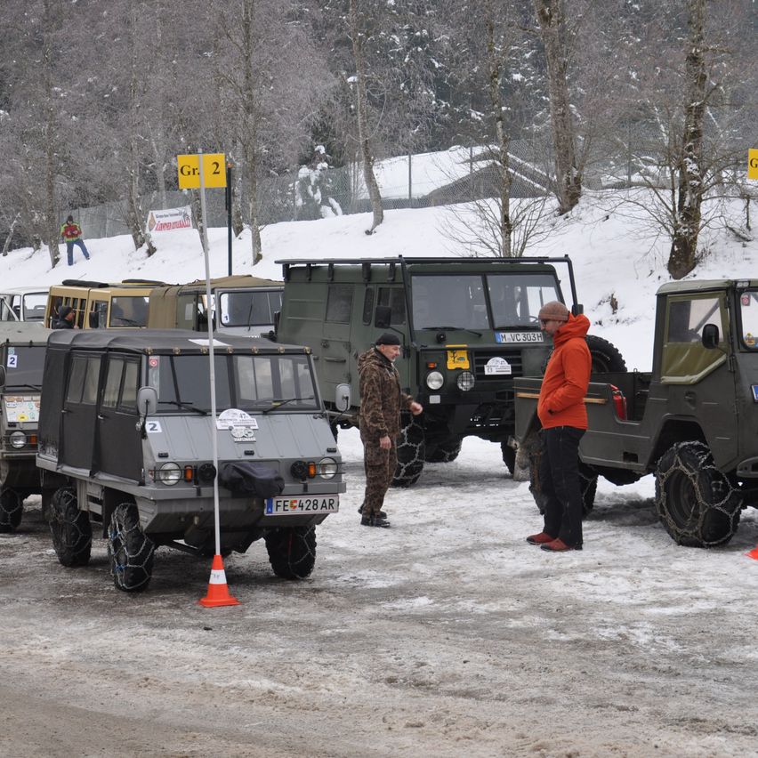 Eine Gruppe militärischer Fahrzeuge steht auf schneebedecktem Boden. Menschen stehen in der Nähe der Fahrzeuge, einer trägt einen orangefarbenen Mantel. Verschneite Bäume und ein Schild mit der Aufschrift Gr. 2 sind sichtbar.