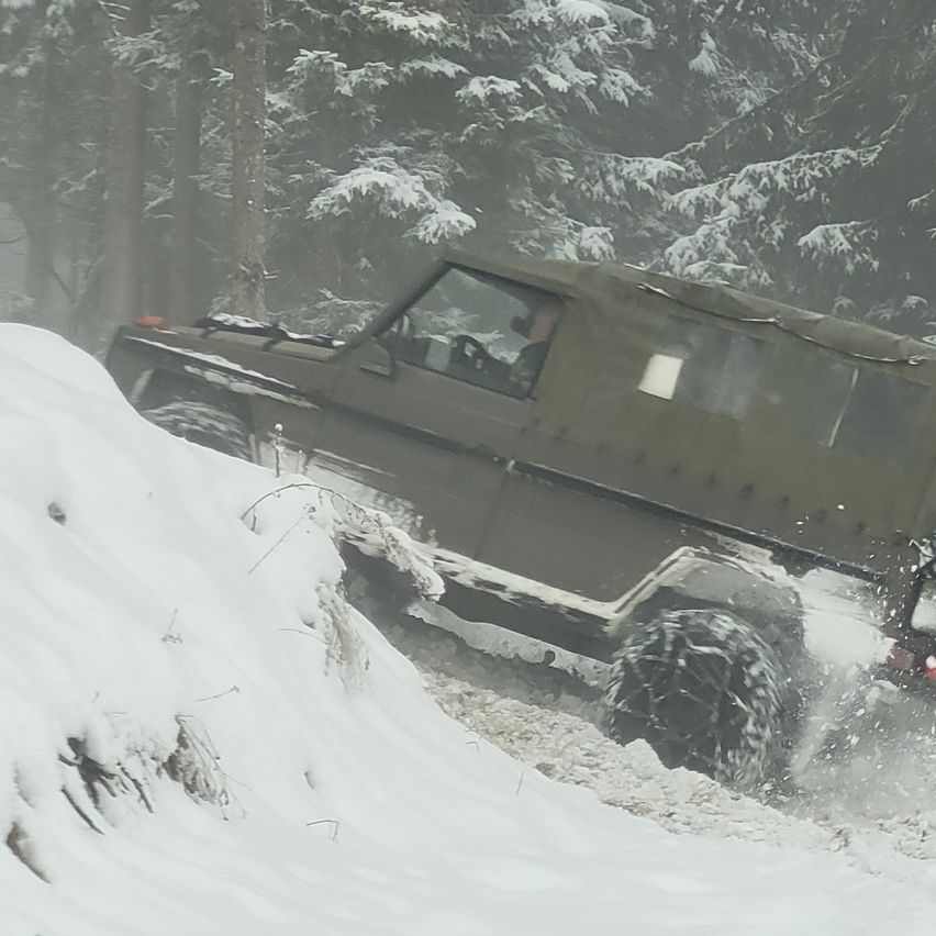 Ein grünes Geländefahrzeug fährt durch einen verschneiten Wald. Das Fahrzeug spritzt Schnee auf, während es sich durch das schneebedeckte Gelände bewegt. Der Fahrer trägt eine Mütze und Handschuhe.