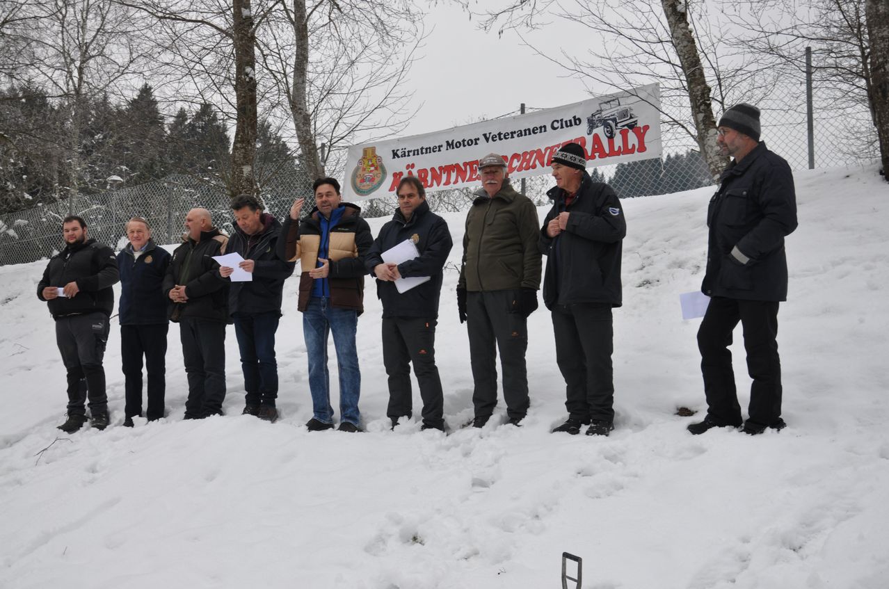 Eine Gruppe von Männern steht in einem verschneiten Feld, hält Papiere in den Händen, und ein Banner mit der Aufschrift 'Kärntner Motor Veteranen Club' befindet sich hinter ihnen.