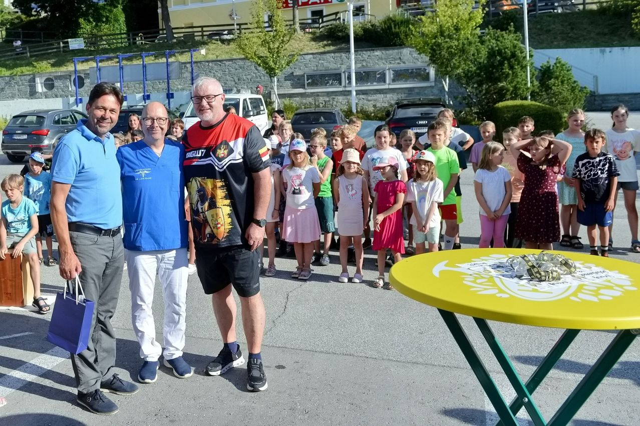 Eine Gruppe von Erwachsenen und Kindern steht in einer Außenumgebung zusammen, mit einem gelben Tisch im Vordergrund. Die Erwachsenen tragen Freizeitkleidung, und die Kinder sind in verschiedenen Outfits gekleidet. Der Hintergrund zeigt eine Steinmauer und ein geparktes Auto.