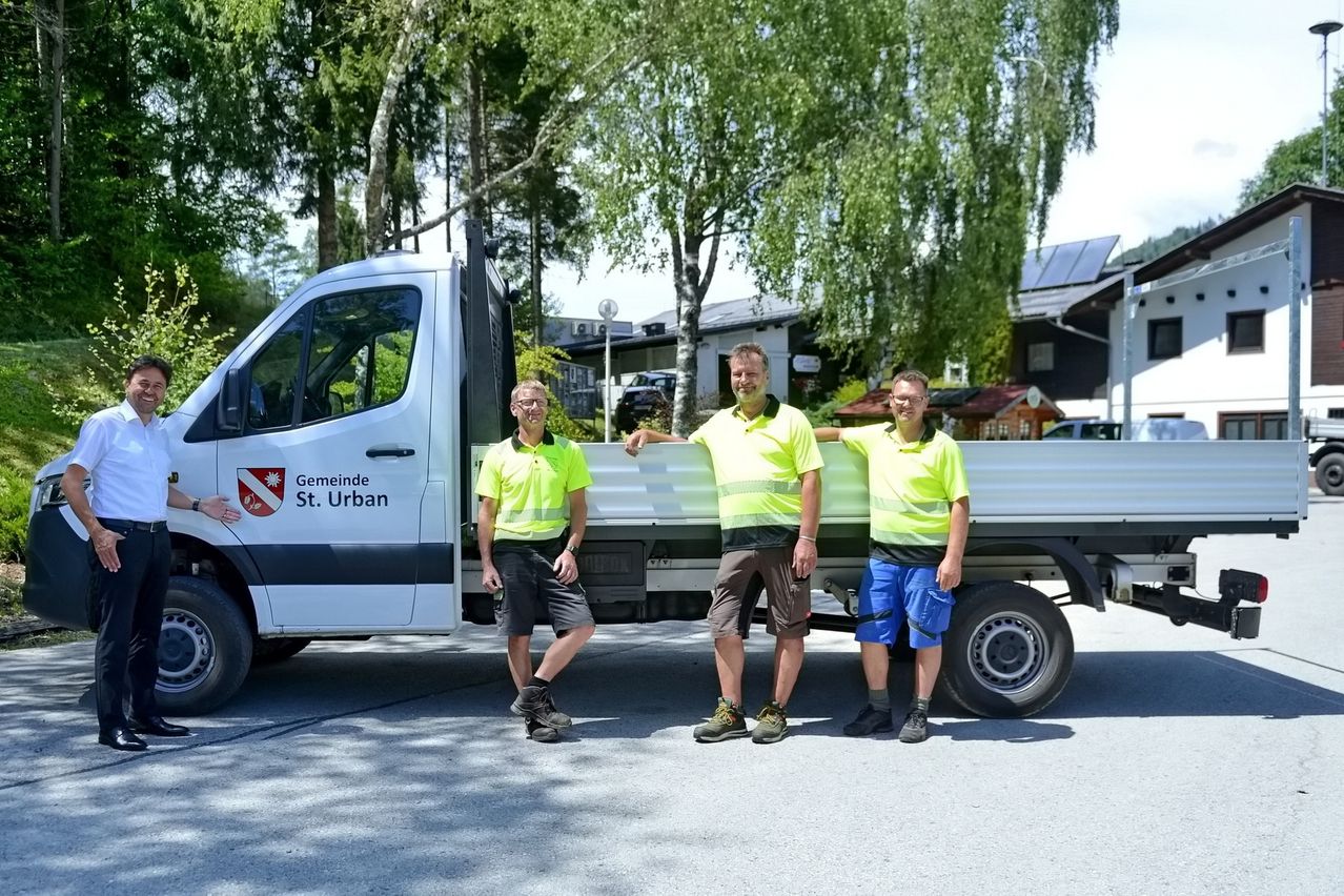 Drei Männer in Sicherheitswesten stehen vor einem weißen Lastwagen mit dem Logo Gemeinde St. Urban. Sie lächeln und posieren für ein Foto. Hinter ihnen sind Bäume und Gebäude.