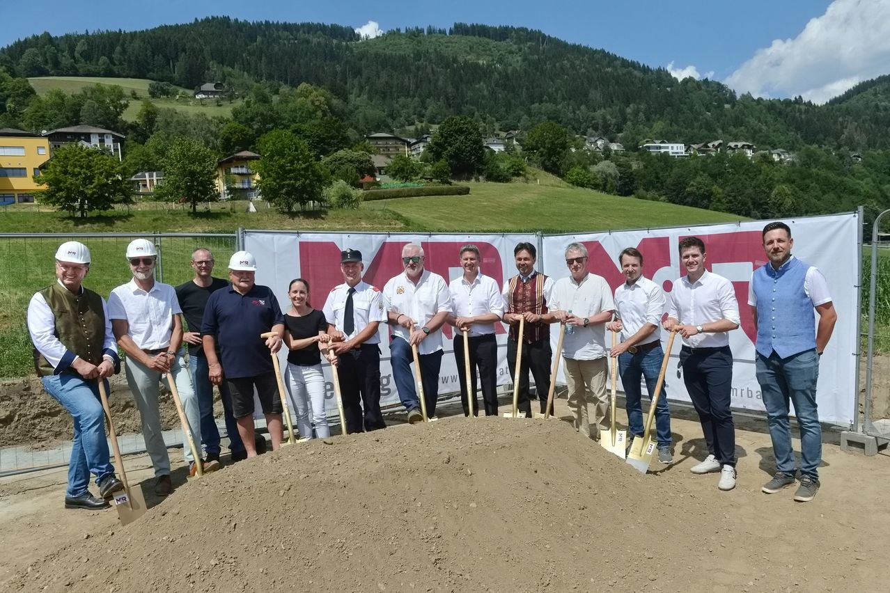 Eine Gruppe von Menschen in formeller Kleidung steht im Kreis und hält Schaufeln, möglicherweise bei einer Grundsteinlegungszeremonie. Hinter ihnen befindet sich ein Banner mit roten Buchstaben und einer bergigen Landschaft im Hintergrund.