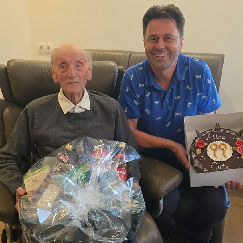 Ein älterer Mann im Rollstuhl lächelt, während er einen Geschenkkorb hält. Neben ihm steht ein Mann, der einen Kuchen mit der Nummer 99 in der Hand hält.