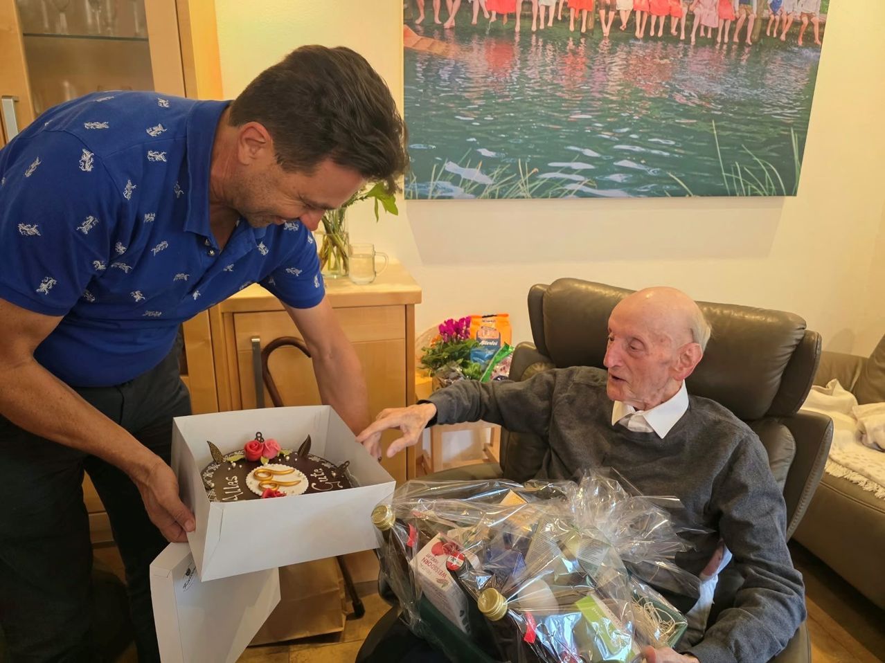 Ein Mann in einem blauen Shirt überreicht einem älteren Mann in einem Stuhl einen Kuchen, mit einem Geschenkkorb neben ihm. Im Hintergrund befindet sich ein Gemälde von Menschen in einem See.