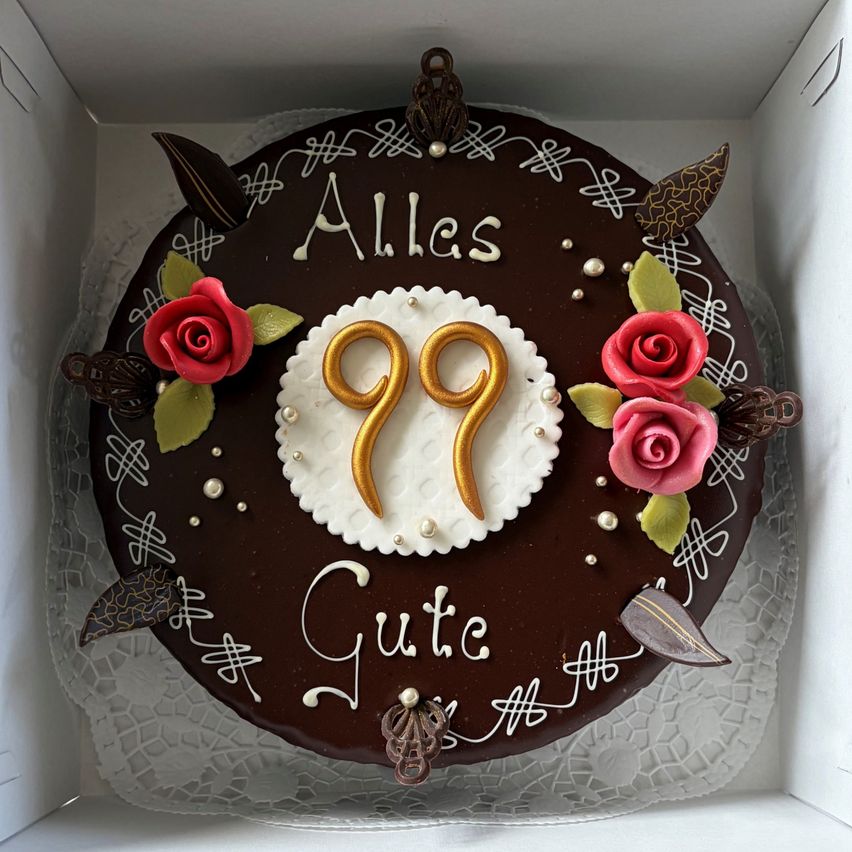 Ein Schokoladenkuchen mit einem weißen Zentrum, verziert mit goldenen Zahlen 99 und roten Rosen, mit den Worten 'Alles Gute' in Weiß auf einem Schokoladenhintergrund.
